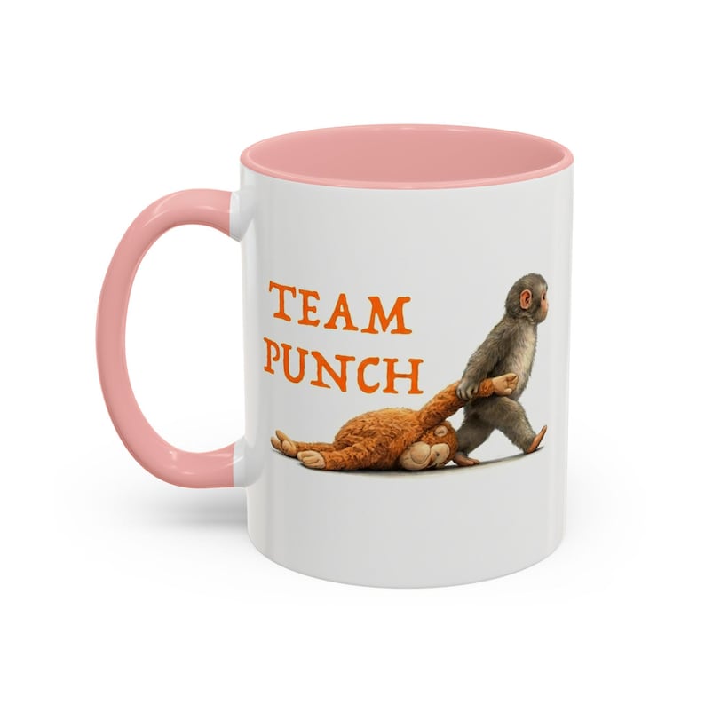 May include: White ceramic mug with a pink interior and handle. The mug features the text "TEAM PUNCH" in orange, along with an illustration of a monkey dragging a stuffed animal. A fun and unique gift.