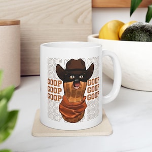 May include: White ceramic mug featuring a cartoon peanut character wearing a cowboy hat, eye mask, and scarf. The mug has the word "GOOP" repeated around the peanut. The mug is on a square coaster.