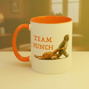 May include: White ceramic mug with an orange interior and handle. The mug features the text "TEAM PUNCH" in orange, along with an illustration of a monkey pulling a stuffed animal. The mug is suitable for hot or cold beverages.