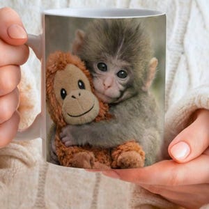 May include: White ceramic mug showing a baby monkey cuddling a stuffed orangutan. The monkey has grey fur and dark eyes, the stuffed toy is brown with a smiling face. The mug is held by a person wearing a white sweater.