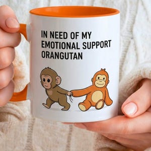 以下が含まれることがあります： オレンジ色の内側とハンドルが付いた白いセラミックマグ。マグには、「IN NEED OF MY EMOTIONAL SUPPORT ORANGUTAN」というテキストと、2匹のオランウータンの漫画イラストが描かれています。オランウータンは茶色とオレンジ色です。