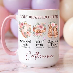 May include: White ceramic mug with a pink handle and rim, featuring the text "GOD'S BLESSED DAUGHTER" and the name "Catherine". The mug is decorated with illustrations of a shield, belt, and sandals, each adorned with flowers and text.