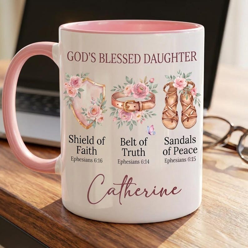May include: White ceramic mug with a pink handle and interior rim. The mug features the text "GOD'S BLESSED DAUGHTER" and illustrations of a shield, belt, and sandals, with the name "Catherine" at the bottom.