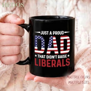 May include: A black ceramic mug featuring the white text JUST A PROUD, the word DAD stylized with the American flag pattern, and the white text THAT DIDN'T RAISE, followed by the red text LIBERALS with two white stars on either side.