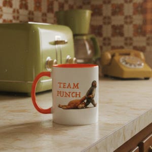 May include: White ceramic mug with an orange interior and handle. The mug features the text "TEAM PUNCH" in orange, along with an illustration of a figure punching another. A retro green toaster and phone are in the background.