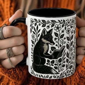 Pode incluir: Uma caneca de cerâmica branca com interior e alça pretos. A caneca apresenta uma ilustração a preto e branco de uma raposa lendo um livro, rodeada por desenhos florais e de folhas. A caneca é segurada por uma pessoa que usa um suéter laranja.
