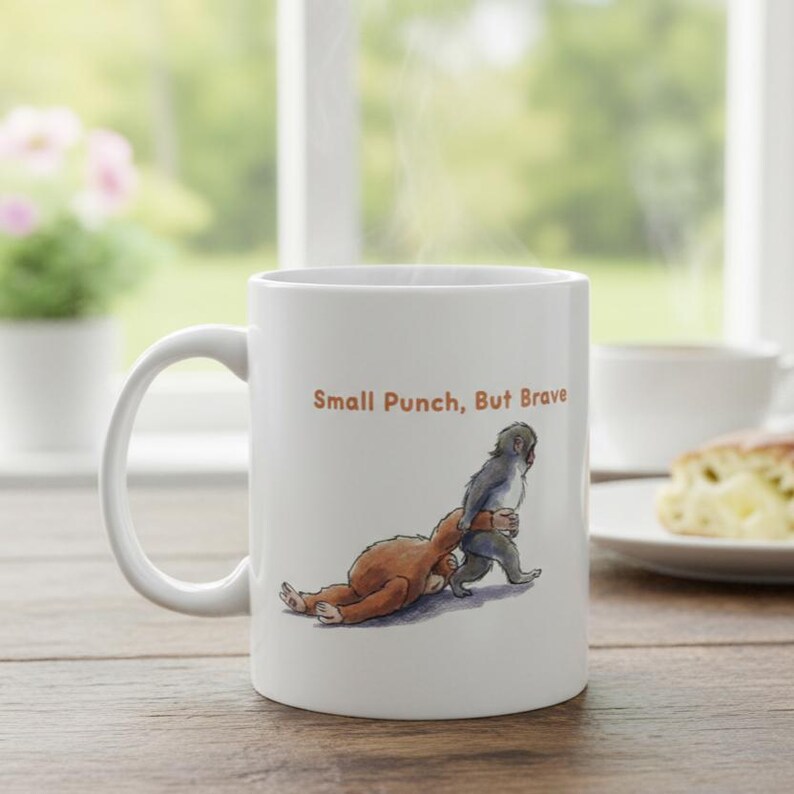 May include: White ceramic mug with a handle, featuring an illustration of two monkeys and the text "Small Punch, But Brave" in orange. The artwork depicts one monkey pulling the other. A plate with food is in the background.