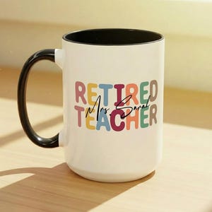 K&ouml;nnte beinhalten: Wei&szlig;er Keramikbecher mit schwarzem Henkel und Innenraum. Der Becher zeigt die Worte "RETIRED Mrs. Sarah TEACHER" in bunten Buchstaben. Ein nachdenkliches Geschenk f&uuml;r einen pensionierten Lehrer.