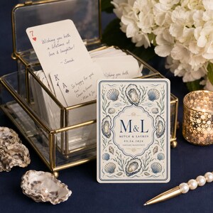 Pu&ograve; includere: Una scatola in vetro e ottone contenente un mazzo di carte con messaggi scritti a mano. Una carta in primo piano presenta un'illustrazione di ostriche blu e crema con il testo M&L, Mitch & Lauren e la data 09.26.2026 stampati sopra.