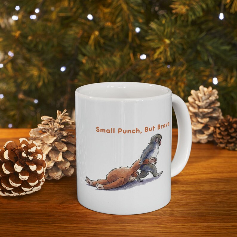 May include: White ceramic mug with a cartoon illustration of two monkeys and the text "Small Punch, But Brave" in brown. The image shows a brown monkey on the ground and a gray monkey standing over it.