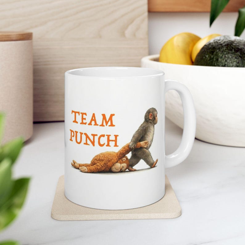 May include: White ceramic mug with the words "TEAM PUNCH" in orange. The mug features a cartoon illustration of a monkey pulling a stuffed animal. The mug is on a beige coaster.