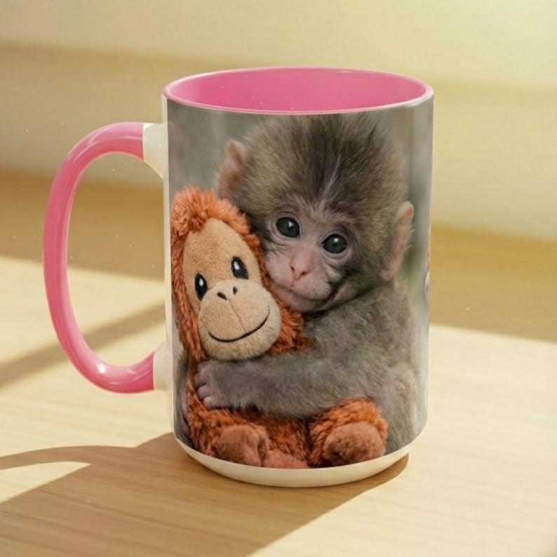 May include: White ceramic mug with a pink interior and handle. The mug features a print of a baby monkey hugging a stuffed orangutan. The monkey has grey fur and the stuffed animal is brown.