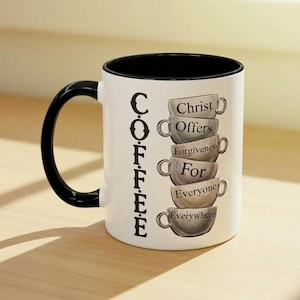 May include: White coffee mug with a black interior and handle. The mug features the word "COFFEE" vertically on the side and a stack of coffee cups with the words "Christ Offers Forgiveness For Everyone Everywhere."