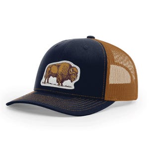 May include: A navy blue trucker hat with a brown mesh back and a curved brim. The hat features a patch with a brown bison illustration. The hat is a classic accessory for outdoor enthusiasts.