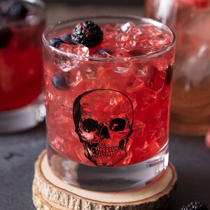 May include: A red cocktail with ice and blackberries in a glass with a skull design. The glass is sitting on a wooden coaster.