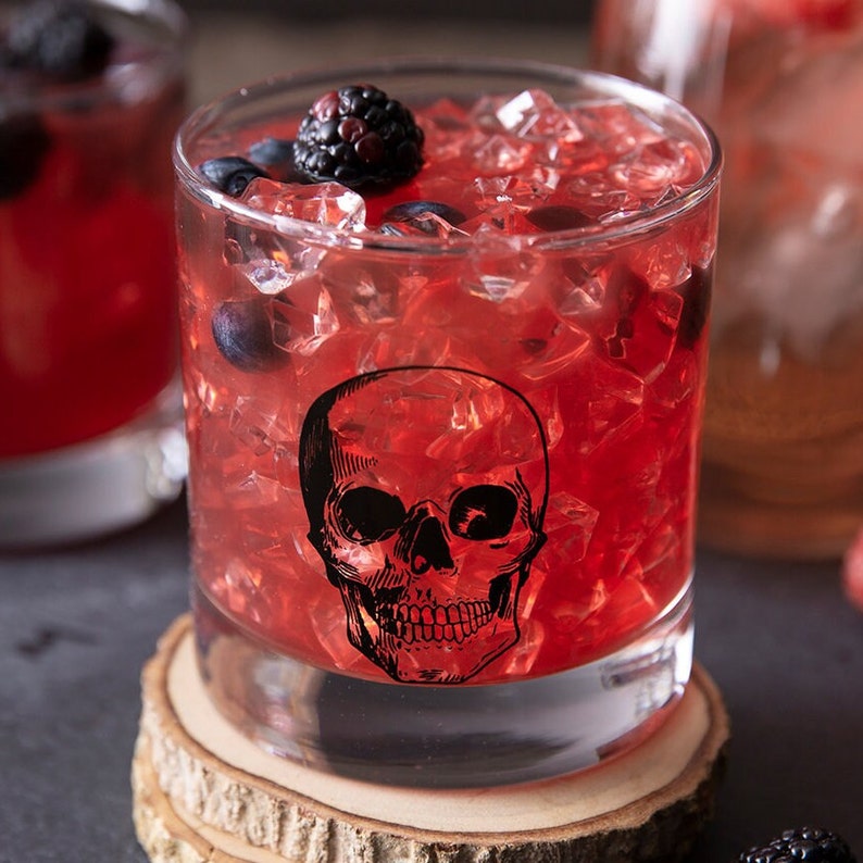 May include: A red cocktail with ice and blackberries in a glass with a skull design. The glass is sitting on a wooden coaster.