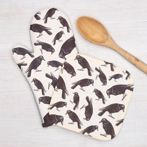 May include: A set of two oven mitts and a pot holder featuring a black and white crow pattern. The oven mitt is a long, single-handed mitt. The pot holder is a square shape.