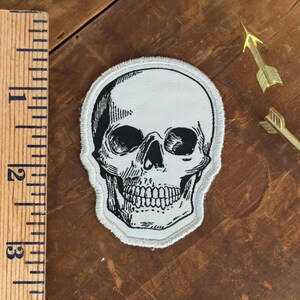 May include: Embroidered patch featuring a detailed black and white skull design. The patch has a light gray background and a stitched border. A wooden ruler shows the patch is approximately 2.5 inches wide.
