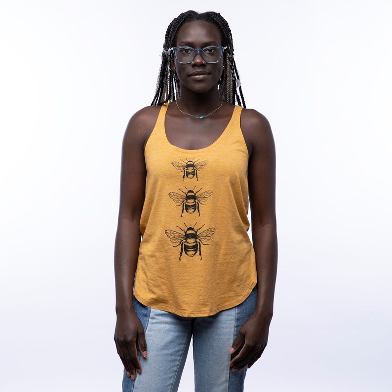 May include: A woman wearing a mustard yellow tank top with a black bee graphic print. The tank top has a scoop neck and a relaxed fit. The woman is wearing blue jeans and has long black braids.