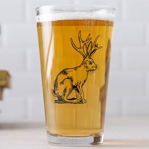 May include: A clear glass pint glass with a black and white illustration of a rabbit with antlers. The glass is filled with a light yellow beer.