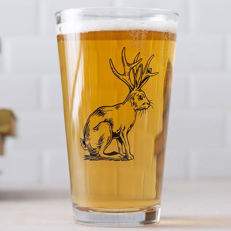 May include: A clear glass pint glass with a black and white illustration of a rabbit with antlers. The glass is filled with a light yellow beer.
