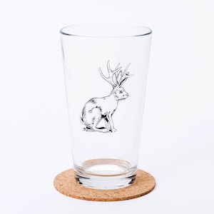 May include: A clear glass pint glass with a black and white illustration of a rabbit with antlers. The glass is sitting on a cork coaster.