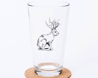 May include: A clear pint glass with a black and white illustration of a jackalope, a mythical creature with the body of a rabbit and antlers. The glass sits on a round, cork coaster. The design is simple and elegant.