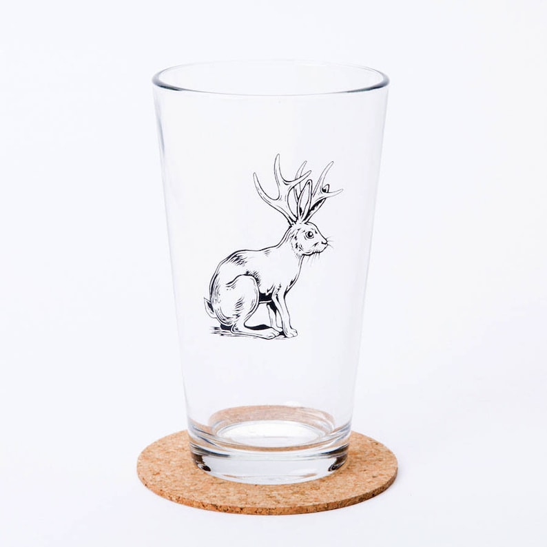 May include: A clear glass pint glass with a black and white illustration of a rabbit with antlers. The glass is sitting on a cork coaster.