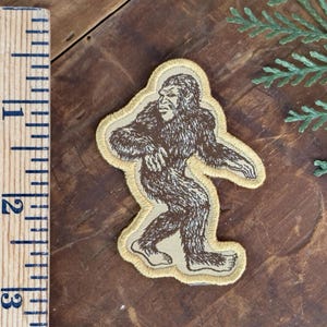 May include: An embroidered patch featuring a brown Bigfoot silhouette on a tan background with a yellow border. A wooden ruler is positioned to the left, showing the patch measures approximately 3 inches in height from top to bottom.