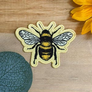 May include: An embroidered patch featuring a bumblebee with a black and yellow striped body, black legs, and white wings with black outlines. The patch has a light yellow border and is shaped to follow the contours of the insect's wings and body.