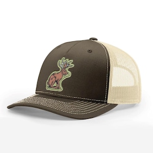 May include: A brown trucker cap with a beige mesh back and a curved peak. The cap features a patch with a brown jackalope illustration with antlers. The peak has white stitching.