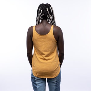 May include: A woman wearing a mustard yellow tank top and blue jeans. She has long black and gray braids.
