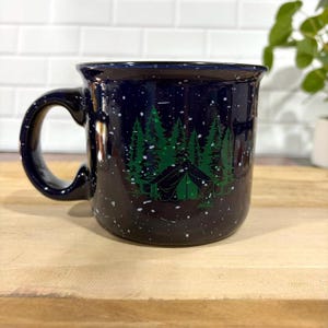 May include: A dark blue ceramic mug with a speckled white finish resembling a starry night sky. The front features a green graphic illustration of a tent nestled among a dense forest of pine trees, creating a nature-inspired outdoor scene.