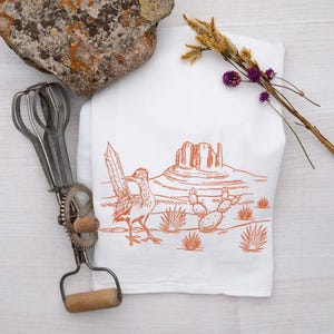 May include: White kitchen towel with an orange illustration of a roadrunner, desert landscape, and cacti. A vintage egg beater and a rock are next to the towel. Dried flowers are on the right.