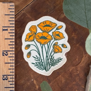 May include: Embroidered patch featuring a bouquet of orange poppies with green stems and leaves on a white background. The patch has a cream-colored border and is approximately 2 inches in height.