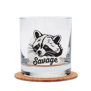 May include: A clear glass tumbler with a black and white raccoon graphic and the word "Savage" in a script font. The glass sits on a round cork coaster. The tumbler is likely for beverages.