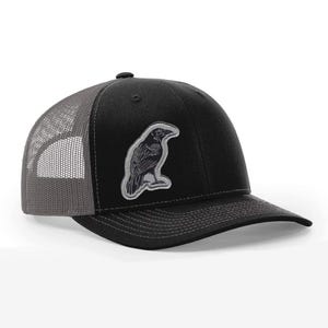 Puede incluir: Gorra de camionero negra con la parte trasera de malla gris y una visera curva. Un parche bordado de cuervo plateado y negro está en la parte delantera. La gorra tiene un cierre a presión ajustable.