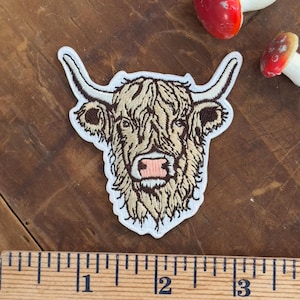 Puede incluir: Parche bordado con la cabeza de una vaca de las Tierras Altas con pelaje marrón y beige, una nariz rosa y cuernos curvados. El parche está sobre una superficie de madera con una regla y dos decoraciones de setas.