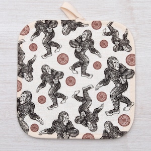 May include: A white cotton pot holder with a repeating pattern of brown footprints and brown circles. The footprints are of a large, hairy creature, resembling a Bigfoot.