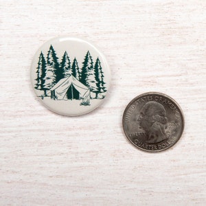 May include: A white button with a green illustration of a tent in a forest. The tent is in the center of the button and is surrounded by five trees. The button is 1 inch in diameter.