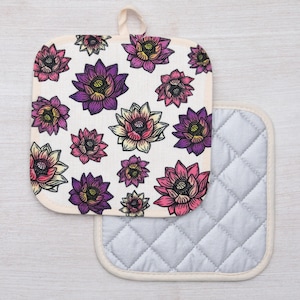 May include: A set of two pot holders. One is a white cotton pot holder with a floral pattern of pink, purple, and yellow flowers. The other is a gray quilted pot holder with a white border.
