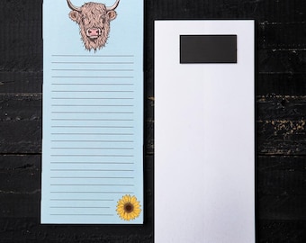 Puede incluir: Un bloc de notas azul claro con una ilustración de una vaca de las Tierras Altas y un girasol. El bloc de notas tiene páginas rayadas. Un bloc de notas blanco con un imán negro está al lado.