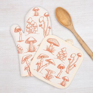 May include: A set of two kitchen oven mitts with a mushroom design. The mitts are made of a white fabric with a red-orange mushroom print. One mitt is a traditional oven mitt shape, and the other is a square pot holder.