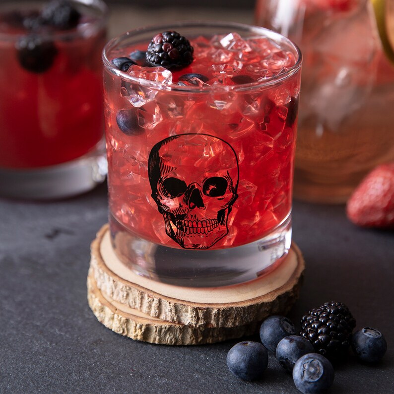 May include: A red cocktail with ice in a glass with a skull design. The glass is sitting on a wooden coaster. There are blueberries and blackberries scattered around the glass.