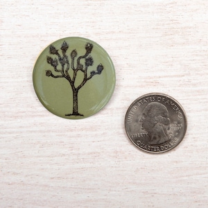 Könnte beinhalten: Ein runder, olivgrüner Knopf mit einer schwarzen Illustration eines Joshua-Baums. Eine US-Viertel-Dollar-Münze liegt neben dem Knopf zum Größenvergleich. Der Knopf hat ein glänzendes Finish und ein naturnahes Design.