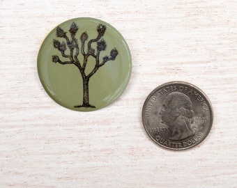 Könnte beinhalten: Ein runder, olivgrüner Knopf mit einer schwarzen Illustration eines Joshua-Baums. Eine US-Viertel-Dollar-Münze liegt neben dem Knopf zum Größenvergleich. Der Knopf hat ein glänzendes Finish und ein naturnahes Design.
