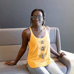 May include: A woman wearing a yellow tank top with a black bee design. She is sitting on a gray couch and has long black braids.