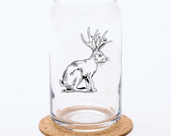 May include: A clear glass tumbler with a cylindrical shape and a curved rim, featuring a black line art illustration of a jackalope, a rabbit with antlers, printed on the front surface. The glass is positioned upright on a round cork coaster.