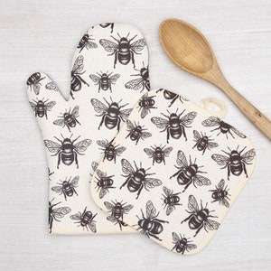 May include: A set of two white cotton oven mitts with a black bee pattern. The mitts are designed for use in the kitchen.