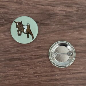 Könnte beinhalten: Zwei runde Ansteckbuttons. Ein Button zeigt eine braune und weiße Ziegenillustration auf hellgrünem Hintergrund. Der andere Button ist silberfarben mit einer Anstecknadel. Die Buttons liegen auf einer braunen Holzoberfläche.
